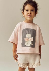 Ljusrosa oversized t-shirt med kaninmotiv och rosett, ihopparad med krämfärgade shorts med spetsdetaljer vid fållen.