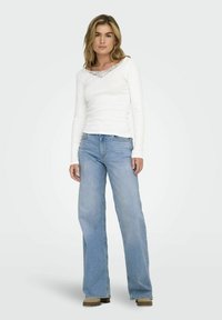 Top alb cu mâneci lungi, cu decolteu din dantelă, asortat cu blugi deschiși la culoare, evazați. Material din bumbac texturat, croi drept și încălțăminte neutră.