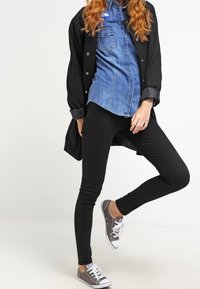 Vrouw in een spijkerblouse, zwarte jas, zwarte skinny broek en grijze sneakers staat met één been gebogen voor een witte achtergrond.