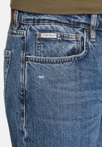 Jeans in denim blu con zone sbiadite e un piccolo etichetta con la scritta "GUESS JEANS." Presentano un classico design a cinque tasche e un rivetto in rame singolo.