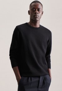 Maglione nero a coste con scollatura tonda e maniche lunghe, abbinato a pantaloni scuri; il modello sta in piedi con le mani nelle tasche.