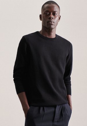 Schwarzer, gerippter Strickpullover mit runden Ausschnitt und langen Ärmeln, kombiniert mit dunklen Hosen; das Modell steht mit den Händen in den Taschen.