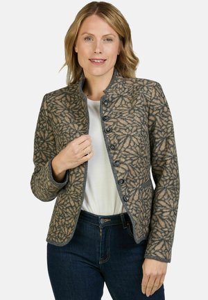 Vrouw draagt een gebloemd jasje met knopen over een wit topje en donkere spijkerbroek, glimlachend, met lichtbruin haar en lichte huid.