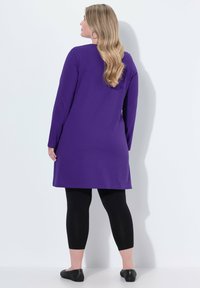 Lila Langarm-Tunika-Kleid aus glattem Stoff, kombiniert mit schwarzen Leggings und flachen Schuhen. Die Haare sind wellig und fallen über die Schultern.