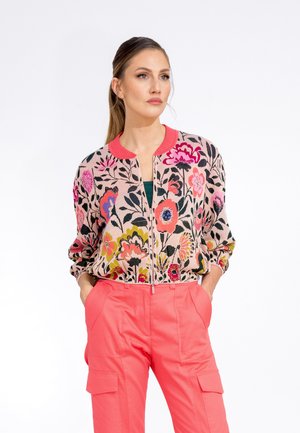 Floral cardigan in pink with contrasting red collar and zip front, featuring vibrant flowers on a textured background. Paired with coral trousers.
