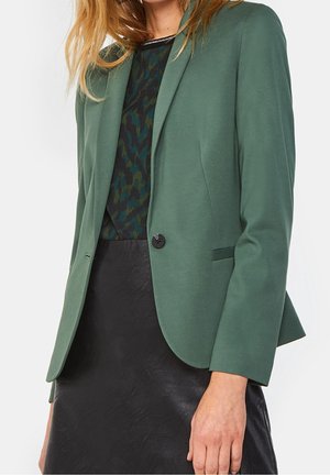 Kvinde iført en grøn en-knaps blazer over en sort mønstret top og sort lædernederdel, stående med hendes højre hånd synlig.
