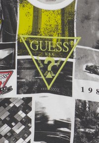 Camiseta gráfica con un collage fotográfico en blanco y negro, acentos amarillos y un destacado logotipo triangular en neón verde que presenta "GUESS U.S.A." y detalles de texto.
