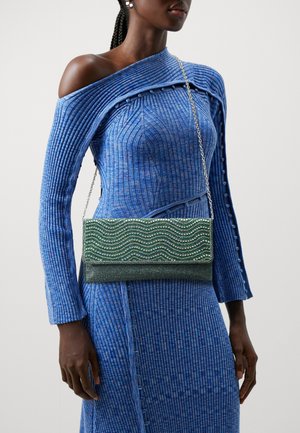 Femme portant une robe bleue côtelée à col bardot et tenant un sac à main vert avec une chaîne et un motif ondulé de strass.