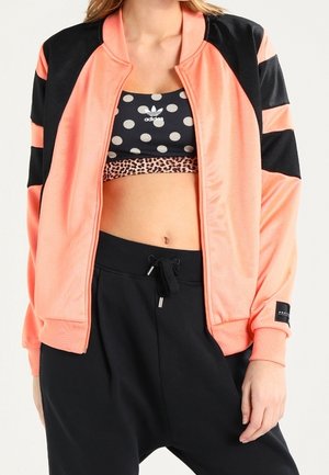 Femme portant une veste zippée pêche et noire, un soutien-gorge de sport noir à pois, et un pantalon noir à cordon, debout avec une main le long du corps.