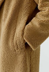 Close-up van de hand van een persoon in de zak van een getextureerde tan sherpa fleece jas met lange mouwen.