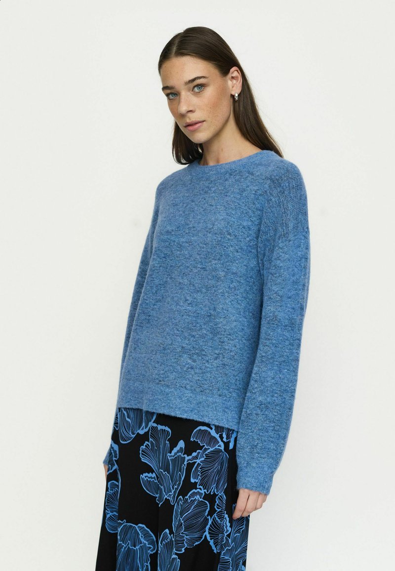 Femme aux cheveux bruns lisses portant un pull bleu et une jupe noire à motif floral bleu se tient devant un fond uni clair.