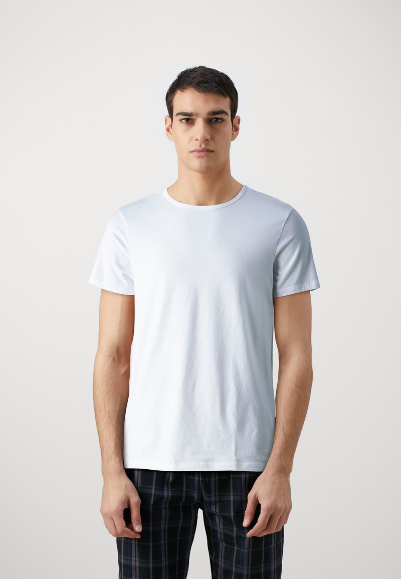 T-shirt en coton bleu clair avec des manches courtes, un col rond et une texture douce. Le modèle le porte avec un pantalon à carreaux noirs.