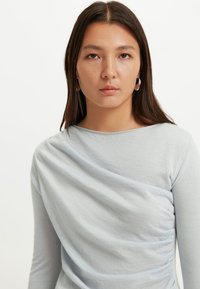 Hellblaues Langarmshirt mit einem fallenden Design, weicher Textur und gerafften Details auf der Vorderseite, die für visuelles Interesse sorgen.