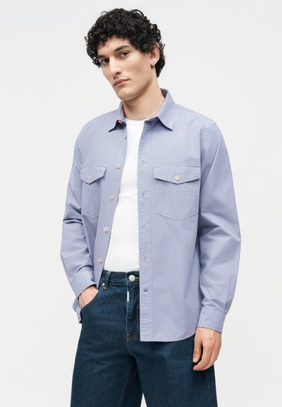 Άνδρας με κυματιστά μαλλιά που φοράει γαλάζιο πουκάμισο πάνω από λευκό μπλουζάκι και σκούρο μπλε τζιν, στέκεται με το ένα χέρι στην τσέπη.