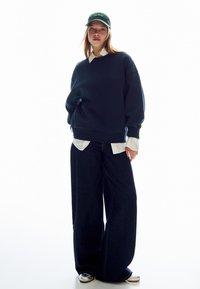 Námořnická mikina s oversize střihem, oblečená přes bílou košilku s límečkem, doplněná širokými tmavými džíny a zelenou čepicí.