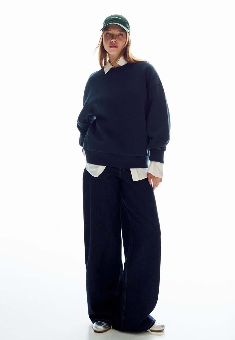 Námořnická mikina s oversize střihem, oblečená přes bílou košilku s límečkem, doplněná širokými tmavými džíny a zelenou čepicí.