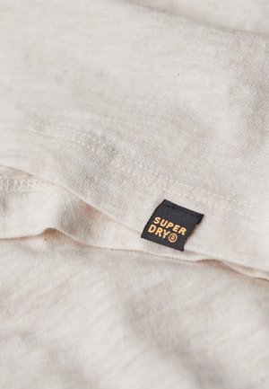 Jasno beżowa bawełniana tkanina o miękkiej strukturze, z czarną metką z napisem "SUPER DRY" w złotym kolorze. Widoczne detale przeszyć.