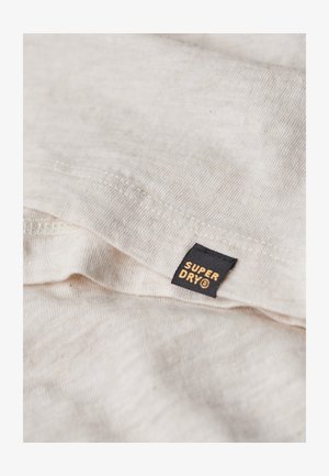 Tessuto di cotone beige chiaro con una consistenza morbida, caratterizzato da un'etichetta nera con "SUPER DRY" in lettere dorate. Dettaglio di cucitura visibile.