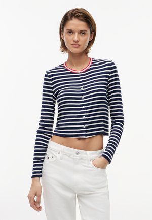 Frau trägt eine marineblaue und weiß gestreifte, langärmelige, kurze Strickjacke mit rotem Besatz und weißen Hosen, posiert mit einer Hand in der Tasche.