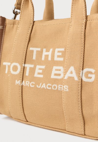 Τσάντα tote από καμβά σε μπεζ χρώμα με λευκό κείμενο που γράφει "THE TOTE BAG MARC JACOBS." Διαθέτει ανθεκτικές λαβές και υφή επιφάνειας.