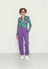 Top de manga longa com padrão verde e roxo, com colarinho, combinado com calças de treino roxas com texto rosa, usado com ténis brancos.