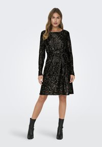 Rochie din catifea neagră cu mâneci lungi, decolteu rotund, talie strânsă și detalii din strasuri sclipitoare. Asortată cu cizme negre până la gleznă.