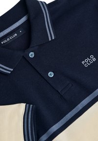 Mörkblå pikétröja med två ljusblå ränder på kragen, utrustad med en ljusbeige sektion och broderad "POLO CLUB"-logotyp.