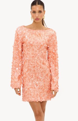 Femme aux cheveux plaqués en arrière, portant de grandes boucles d'oreilles florales et une mini-robe manches longues à sequins pêche, debout sur fond blanc.
