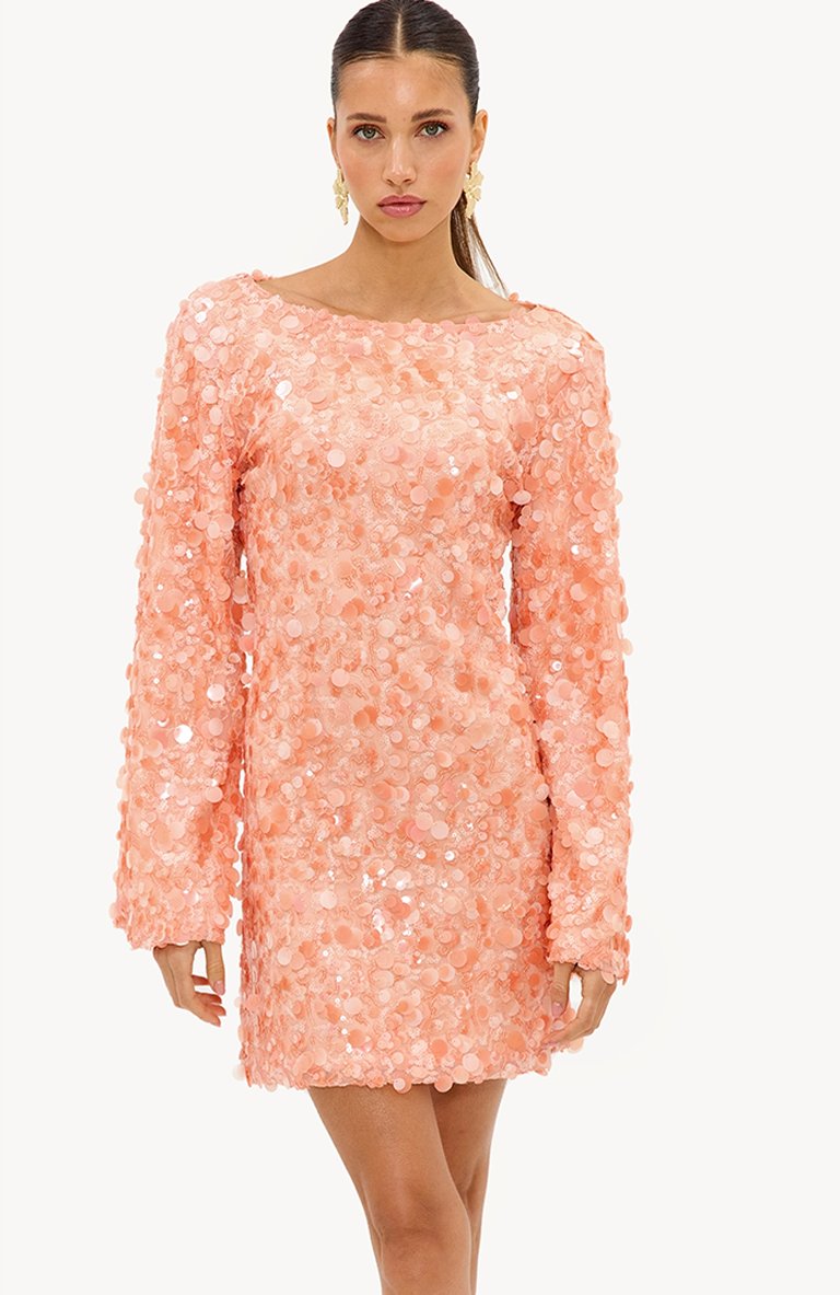 Femme aux cheveux plaqués en arrière, portant de grandes boucles d'oreilles florales et une mini-robe manches longues à sequins pêche, debout sur fond blanc.