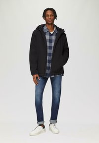 Zwarte hoodie-jack over een blauw en grijs geruit shirt, gecombineerd met slim fit blauwe jeans en witte sneakers met zwarte accenten.