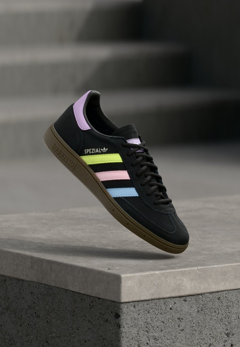 Baskets Adidas Spezial noires avec bandes jaune pastel, rose et bleue, languette de talon lavande et semelle marron, flottant au-dessus d’un rebord en béton.