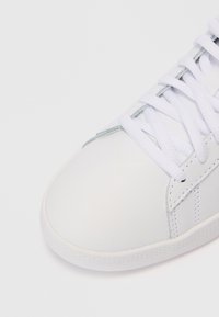 Sapatilha branca com atacadores brancos, costuras visíveis e uma borda de sola texturizada sobre um fundo branco.