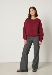 Burgundi spalvos megzto džemperio su nuleistais pečiais ir siuvinėtu logotipu derinys su pilkais platėjančiais keliais ir rudais aulinukais.