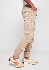 Pantalons cargo beiges avec taille élastique, poches latérales et arrière. Portés avec des chaussures blanches ornées d'accents orange et de semelles noires.