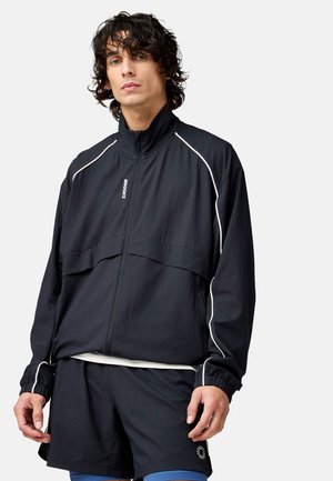 Ung mand med krøllet hår iført en sort træningsjakke med lynlås og hvide detaljer samt matchende shorts med et lille logo.