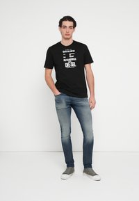 Czarny bawełniany t-shirt z białym nadrukiem, zestawiony z wyblakłymi niebieskimi jeansami o wąskim kroju i szarymi sneakersami. Model stoi przodem.