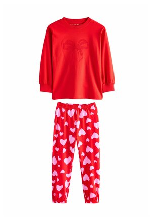Top roșu cu mânecă lungă și design cu fundiță, purtat cu pantaloni roșii cu model de inimioare albe. Țesătura pare moale și confortabilă.