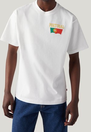 Mies, der ein weißes T-Shirt mit dem Text "Portugal" und dem Wappen der portugiesischen Flagge auf der oberen linken Brust trägt, kombiniert mit blauen Jeans.
