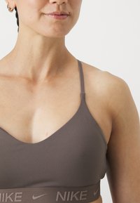 Barna sportmelltartó vékony pántokkal, V-nyakkivágással és struktúrált derékrésszel, melyen a Nike logó kontrasztos árnyalatban látható.