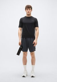 Tricou sport negru cu mâneci scurte asortat cu pantaloni scurți gri închis. Pantofi sport albi și o sticlă de apă neagră în mână. Textură fină.