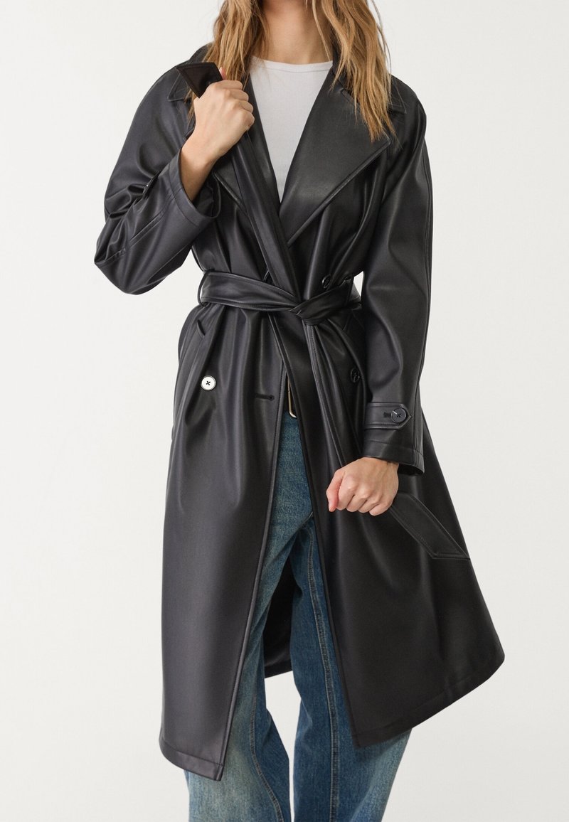Femme portant un trench-coat en cuir noir ceinturé sur un top blanc et un jean bleu, ajustant le col d'une main.