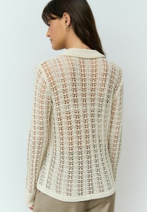 Háčkovaný cardigan v off-white barvě s texturovaným, otevřeným pleteným designem. Obsahuje klasický límec a dlouhé rukávy, s mírně zakřiveným lemem.