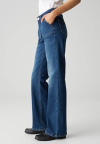 Jeans a zampa d'elefante in denim blu con tasche anteriori e posteriori. Presentano un girovita alto e dettagli di cucitura sottili lungo le cuciture laterali.