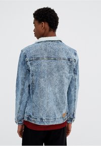 Joven con pelo rizado corto, llevando una chaqueta de mezclilla azul con cuello de forro de lana blanca, una camisa de rayas rojas y pantalones negros, de espaldas.