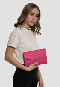 Borsa a mano in pelle rosa con superficie texture e chiusura a patta, tenuta in una mano contro uno sfondo neutro. Dettaglio logo dorato presente.