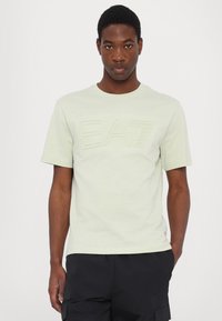 T-shirt en coton vert clair avec un grand logo en relief "EA7". Manches courtes et col rond, avec une texture lisse et une coupe décontractée.
