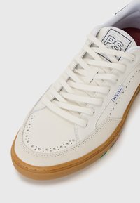 Witte leren sneakers met een geperforeerde neus, platte veters en een tan rubberen zool met veelkleurige accenten aan de zijkant.