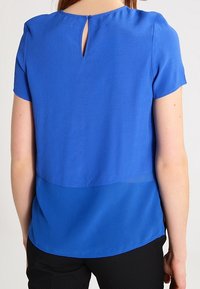 Top blu corto a maniche corte con dettaglio a oblò sul retro e design a strati, con una texture liscia e un pannello inferiore leggermente trasparente.