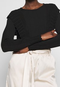 Haut noir à manches longues avec texture côtelée et accents à volants sur les épaules, associé à un pantalon en satin de couleur crème, noué à la taille.