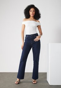 Witte off-shoulder top, donkerblauwe hoge taille flare jeans, zwarte hak sandalen, natuurlijk krullend haar, minimalistische studio achtergrond.